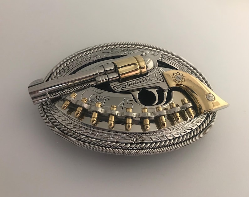 May include: An oval-shaped silver and gold belt buckle featuring a detailed revolver design. The gun is silver with gold accents, and the buckle includes the text "COLT 45". The buckle has a rope-like border.