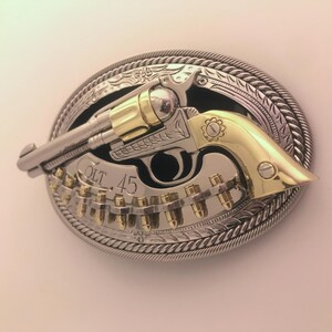 May include: An oval-shaped belt buckle featuring a silver and gold revolver design. The buckle has a silver rope-like border and the text "COLT 45". The revolver is silver with gold accents and is set against a silver background.