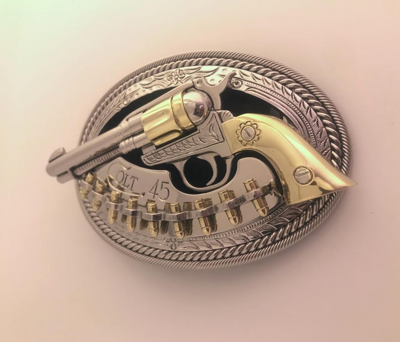 May include: An oval-shaped belt buckle featuring a silver and gold revolver design. The buckle has a silver rope-like border and the text "COLT 45". The revolver is silver with gold accents and is set against a silver background.