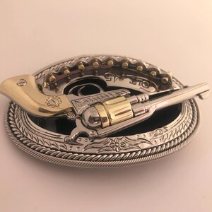 May include: A silver and gold-tone metal belt buckle featuring a revolver design. The buckle has an oval shape with a detailed rope-like border and a black center. The revolver has gold-tone accents and the text "COLT 45".