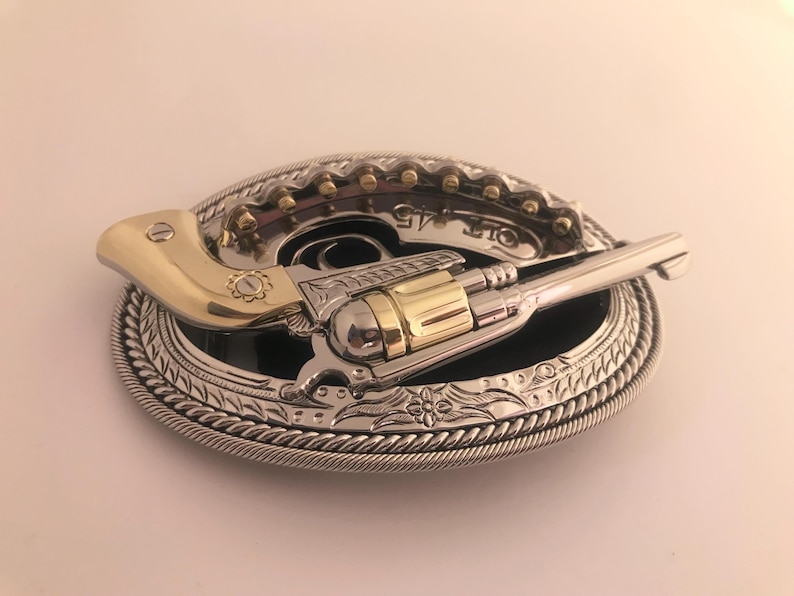 May include: A silver and gold-tone metal belt buckle featuring a revolver design. The buckle has an oval shape with a detailed rope-like border and a black center. The revolver has gold-tone accents and the text "COLT 45".