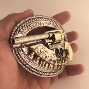 May include: Silver and gold-toned belt buckle featuring a revolver design. The buckle has a detailed border and includes the text "COLT .45". The revolver is depicted with bullets.