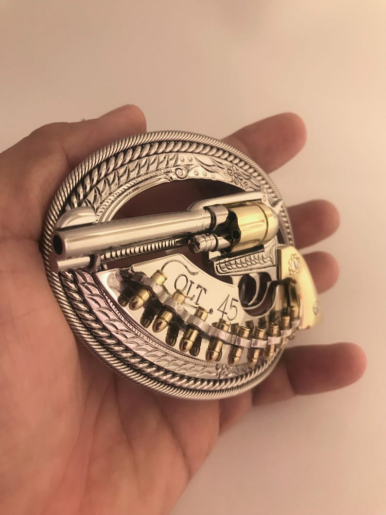 May include: Silver and gold-toned belt buckle featuring a revolver design. The buckle has a detailed border and includes the text "COLT .45". The revolver is depicted with bullets.