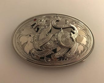May include: A silver belt buckle featuring an elaborate design of a mythical creature. The buckle has a red stone set in the centre.