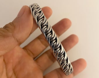 May include: A silver-colored bracelet with a woven chain design. The bracelet is made of metal and has a polished finish. The links are tightly interlocked, creating a solid, textured appearance. The bracelet is approximately 0.75 inches wide.