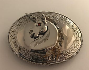 May include: Silver belt buckle featuring an intricate design of a unicorn's head with a red gemstone eye. The buckle has a rope-like border and a textured background.
