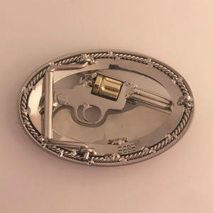 May include: Oval silver-tone belt buckle featuring a detailed revolver design with a gold-colored cylinder. The buckle has a rope-like border and a silver-tone metal clasp. The overall design is Western-themed.