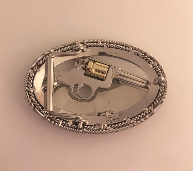 May include: Oval silver-tone belt buckle featuring a detailed revolver design with a gold-colored cylinder. The buckle has a rope-like border and a silver-tone metal clasp. The overall design is Western-themed.