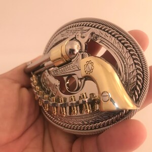 May include: A silver and gold-tone metal belt buckle featuring a detailed revolver design. The buckle has a decorative border with a rope-like edge and engraved patterns. The revolver has gold accents and a row of bullets.