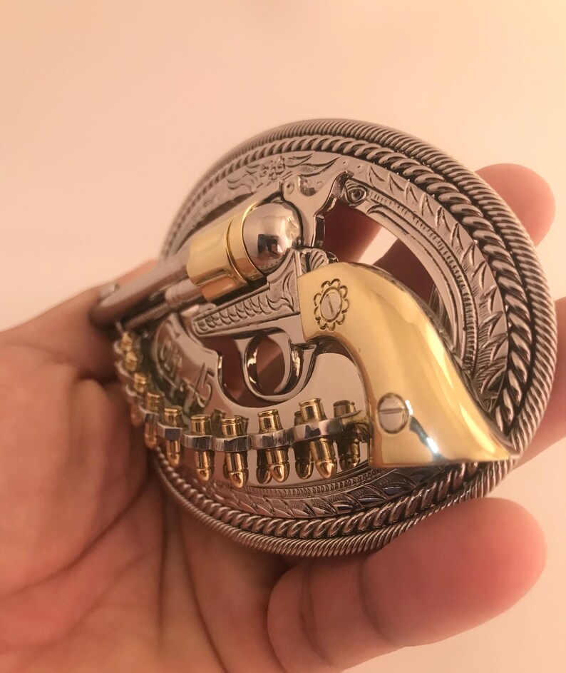 May include: A silver and gold-tone metal belt buckle featuring a detailed revolver design. The buckle has a decorative border with a rope-like edge and engraved patterns. The revolver has gold accents and a row of bullets.
