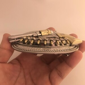 May include: A silver and gold-toned metal belt buckle in the shape of a revolver. The buckle features a detailed design with bullet-shaped accents and a decorative border. The revolver's handle and cylinder are gold-colored.