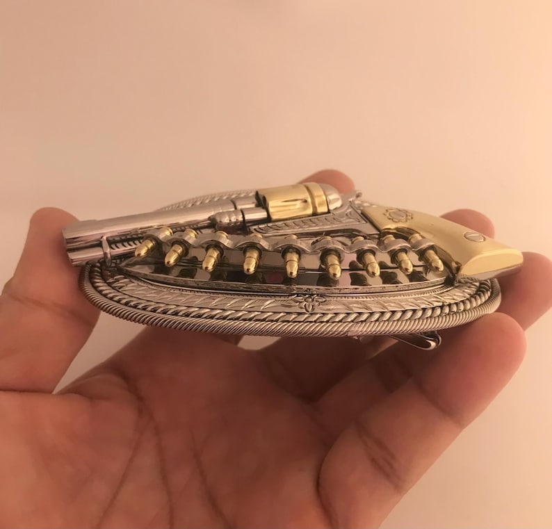 May include: A silver and gold-toned metal belt buckle in the shape of a revolver. The buckle features a detailed design with bullet-shaped accents and a decorative border. The revolver's handle and cylinder are gold-colored.