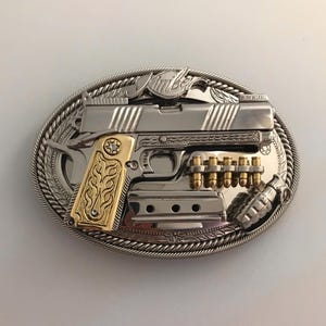 Puede incluir: Hebilla de cinturón de metal plateado y dorado con diseño de pistola. La hebilla tiene un borde en forma de cuerda e incluye un cargador, balas y una granada. La palabra "Kimber" es visible en la parte superior.