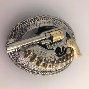 May include: A silver and gold-tone metal belt buckle featuring a revolver design. The buckle has a detailed rope-like border and the text "COLT 45". The revolver and bullets are gold-colored.