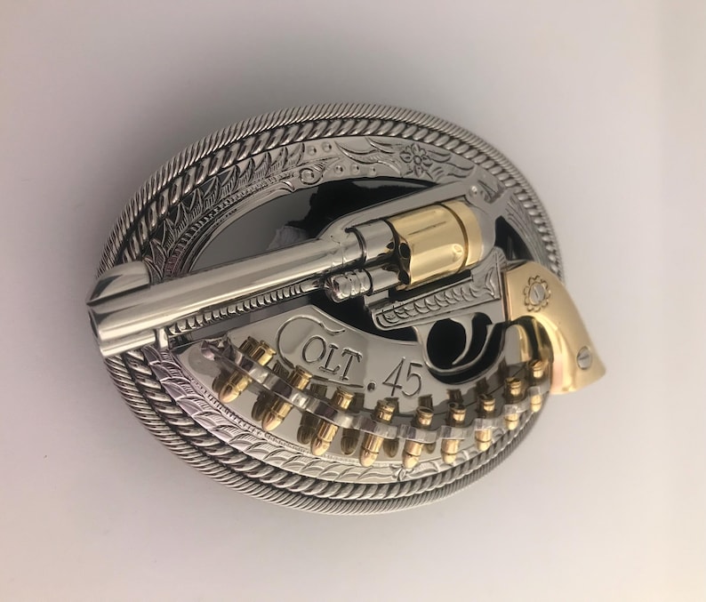 May include: A silver and gold-tone metal belt buckle featuring a revolver design. The buckle has a detailed rope-like border and the text "COLT 45". The revolver and bullets are gold-colored.