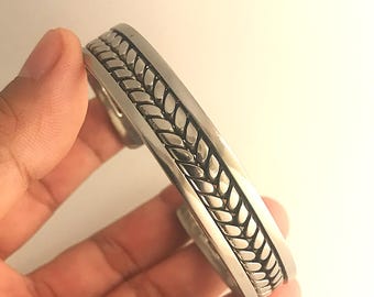 May include: A silver cuff bracelet with a braided design. The bracelet is open ended and has a smooth finish.