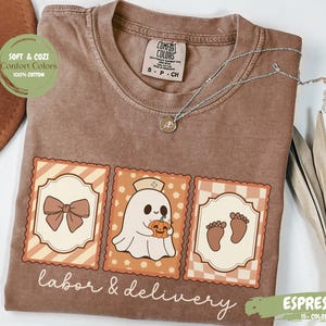 May include: Espresso-colored t-shirt with the words "labor & delivery" and three illustrated stamps. The stamps feature a bow, a ghost holding a pumpkin, and baby footprints. A silver necklace rests on the shirt.