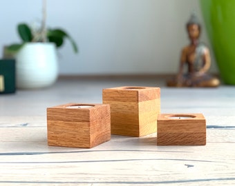 May include: Three wooden candle holders, each with a square shape and a natural wood finish. The candle holders are arranged in a row on a light wood surface.