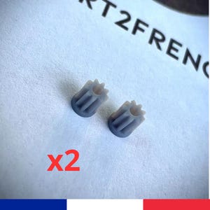 May include: Two small, grey, gear-shaped components are displayed on a textured, off-white surface. The components have a cylindrical body with a star-like top and vertical grooves. The text "x2" is in red below the components.