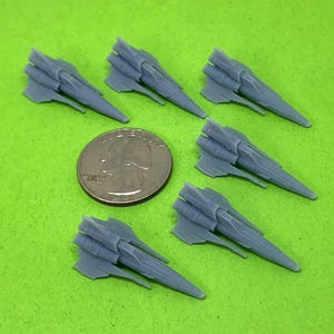 May include: Six small, gray spaceship models are arranged on a green surface, next to a quarter coin for size comparison. The spaceships have a detailed design with wings and multiple engines, suggesting a science fiction theme.