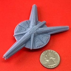 May include: A gray 3D printed model of a spaceship with three arms extending from a central circular base. The model is about the size of a quarter.