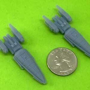 May include: Two gray plastic miniature spaceships, one slightly larger than the other, are shown on a green surface. A silver US quarter is included for scale.