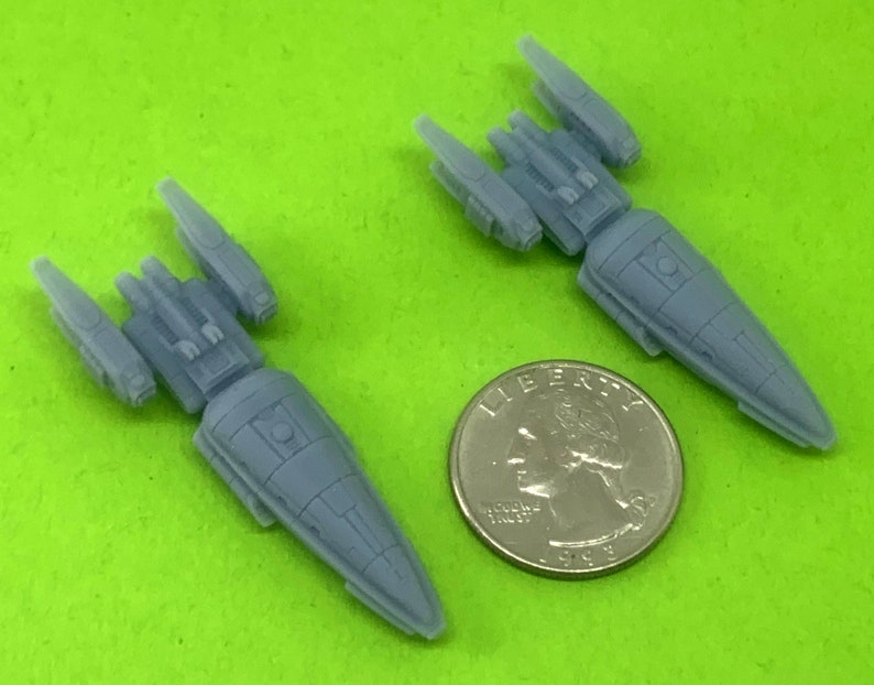 May include: Two gray plastic miniature spaceships, one slightly larger than the other, are shown on a green surface. A silver US quarter is included for scale.