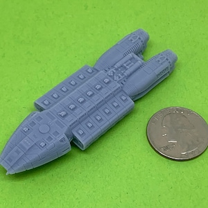 May include: A light gray model of a spaceship with intricate details, next to a U.S. quarter coin on a green surface. The spaceship has a rectangular body with multiple rectangular and circular features.