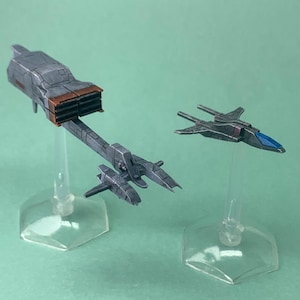 May include: Two miniature gray spaceship models. One is a larger, detailed spacecraft with a brown accent, and the other is a smaller fighter jet with a blue cockpit. Both are mounted on clear stands, set against a green background.