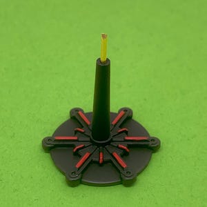 May include: A black and red decorative stand with a central black post and a circular base. The base features red accents radiating from the center. A yellow, cylindrical object extends from the top of the post.