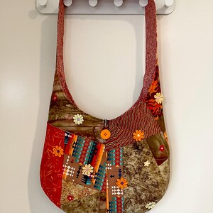May include: A patchwork shoulder bag with a long strap. The bag features a variety of fabrics in shades of red, brown, orange, and teal, with floral and geometric patterns. A large orange button secures the bag's opening.