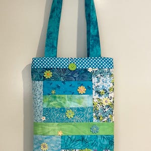 May include: A patchwork tote bag in shades of blue and green, with floral and patterned fabrics. The bag has turquoise handles and a gingham top. Decorative flower-shaped buttons are sewn onto the front.