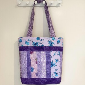 May include: A quilted tote bag in shades of purple, featuring floral and patterned fabric panels. The bag has a top band with blue floral accents and a matching patterned strap. The design includes various shades of purple and lavender.