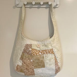 May include: A cream-coloured quilted shoulder bag with a patchwork design. The bag features fabrics in cream, tan, and brown, with floral and paisley patterns. Small white flower embellishments and a wooden button add detail.
