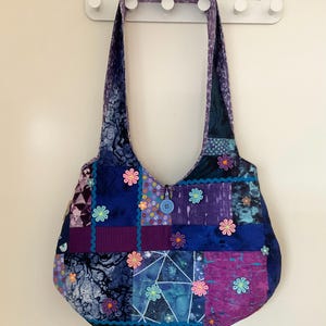 May include: A patchwork shoulder bag in shades of blue and purple, with a button closure and floral appliqués. The bag's strap matches the patterned fabric of the bag. The bag's dimensions are approximately 30 cm wide.