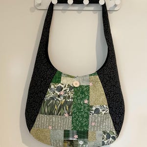 May include: A patchwork shoulder bag with black floral sides and strap. The centre features green and grey patterned fabrics, small pink flower embellishments, and a wooden button closure. The bag hangs from a white wall hook. The bag is approximately 40 cm wide.