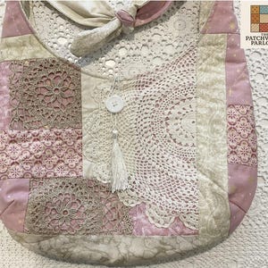 May include: A patchwork shoulder bag with a cream lace doily and a white tassel. The bag features pink and cream fabrics, including floral and star patterns. It has a knotted strap and a button closure. The bag is handmade.