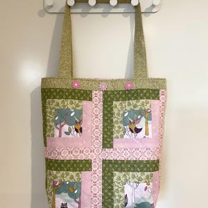 May include: A patchwork tote bag in green and pink. The bag features owl and tree designs within the patchwork. It has two long handles and is decorated with pink flower accents. The bag is suitable for carrying items.