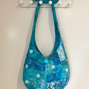 May include: A patchwork shoulder bag in teal and blue hues, decorated with white floral patterns. The bag has a button fastening and a long strap that ties at the top. The bag is hanging on a white wall.