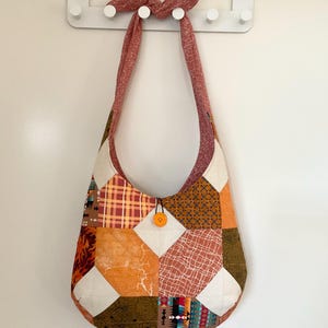 May include: A patchwork shoulder bag with a curved shape and a long strap. The bag features a variety of fabric patterns in shades of orange, brown, and cream, with a button closure. The strap is a reddish-brown colour.