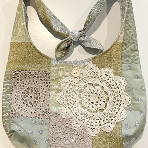 May include: A patchwork shoulder bag in shades of green, beige, and cream. The bag features a large, cream-coloured doily and a wooden button closure. The strap is tied in a knot. The bag is made of various patterned fabrics.