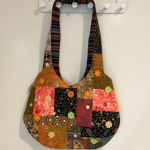 May include: A patchwork shoulder bag in various colours and patterns, including orange, pink, green, and black. The bag has a button closure and is decorated with small flower appliqués. The strap is striped.