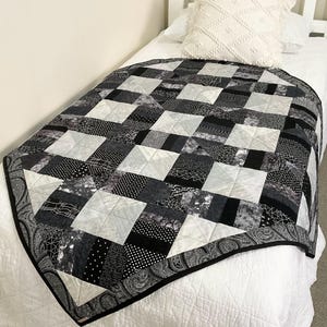 May include: A monochrome patchwork quilt in black, white, and grey, with a paisley border. The quilt displays a geometric pattern of squares and rectangles in various shades. It is placed on a white bed.
