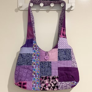 May include: A patchwork shoulder bag in shades of purple. The bag has a button closure and long straps. The design includes floral, polka dot, and geometric patterns. The bag is hanging on a white wall.
