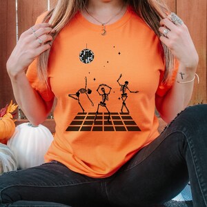 May include: Orange t-shirt featuring a black graphic of dancing skeletons on a checkered dance floor, with a disco ball and stars. The word "Orange" is printed at the bottom. A fun, Halloween-themed design.