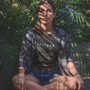 Puede incluir: Una mujer se sienta con las piernas cruzadas en un exuberante jard&iacute;n verde, vistiendo una camiseta negra con la palabra "INSIDER" impresa en ella, unos pantalones cortos de mezclilla azul y una bata de kimono estampada. La luz del sol se filtra a trav&eacute;s de las hojas, proyectando sombras sobre ella.