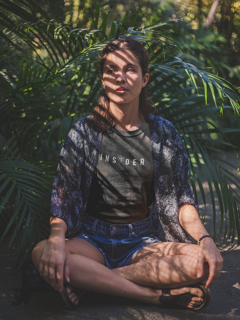 Puede incluir: Una mujer se sienta con las piernas cruzadas en un exuberante jard&iacute;n verde, vistiendo una camiseta negra con la palabra "INSIDER" impresa en ella, unos pantalones cortos de mezclilla azul y una bata de kimono estampada. La luz del sol se filtra a trav&eacute;s de las hojas, proyectando sombras sobre ella.