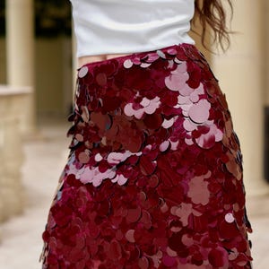 May include: A short skirt covered in overlapping circular burgundy sequins of varying sizes. The skirt features a high-waisted design and is paired with a plain white top. The sequins create a dense, reflective, and textured surface across the garment.