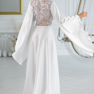 May include: A white maxi dress with long sleeves and a floral lace back detail. The dress has a flowing skirt and a relaxed fit.