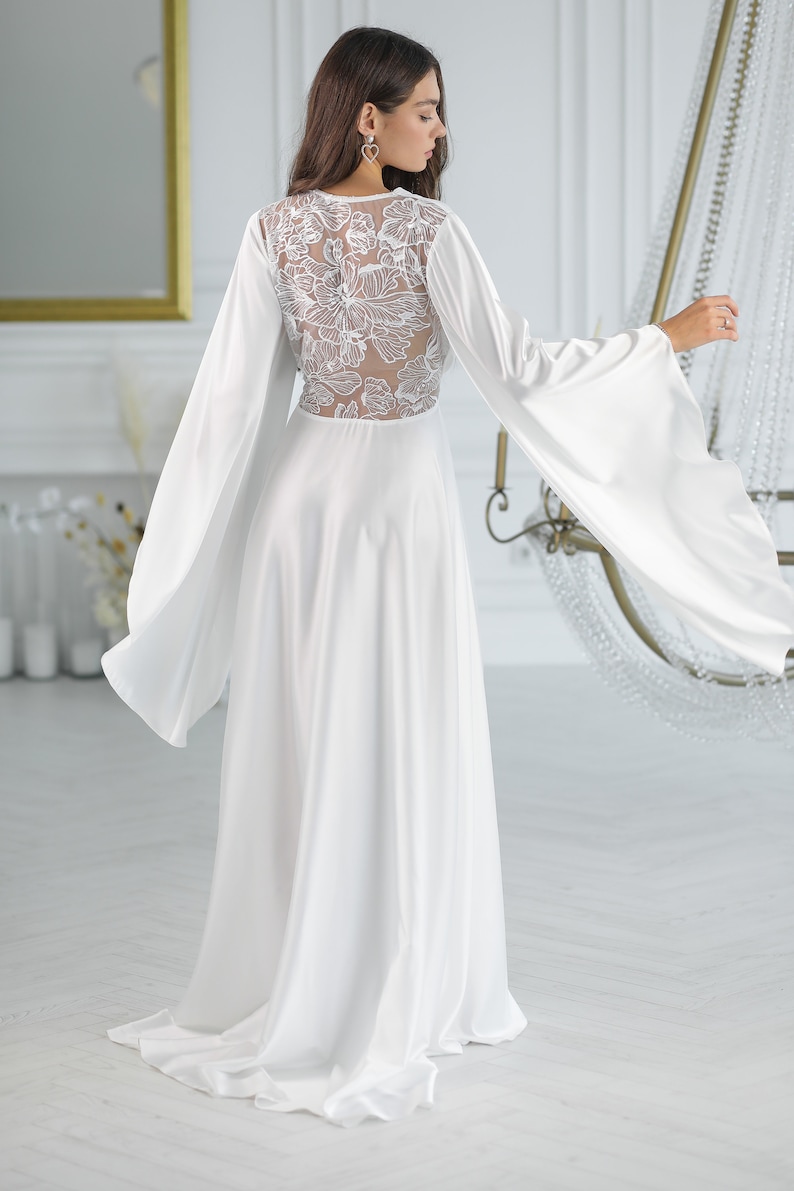 May include: A white maxi dress with long sleeves and a floral lace back detail. The dress has a flowing skirt and a relaxed fit.